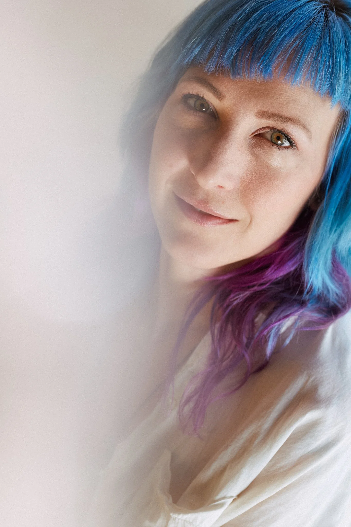 Portrait d'une femme avec des cheveux mi-longs peints en bleu, violet et rose, souriant légèrement, regardant vers la caméra.